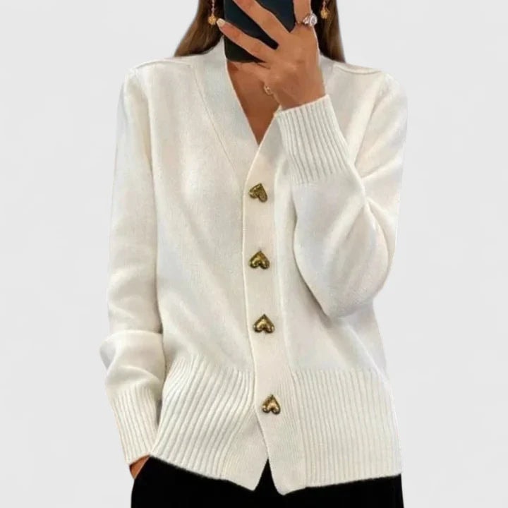 White V-neck cardigan with gold heart-shaped buttons, long sleeves, and ribbed trim, paired with black trousers.