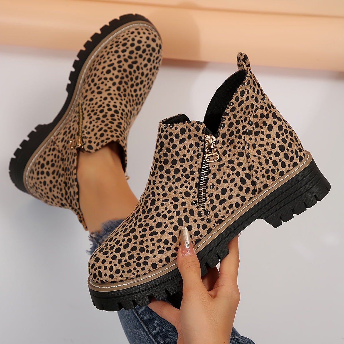 Leopard-print ankle boot held in hand beside its matching boot, with the side zip and lug sole visible.