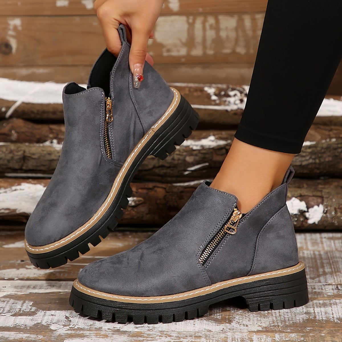 Gray ankle boot held by its pull tab beside the matching boot, displaying the side zip and chunky tread.