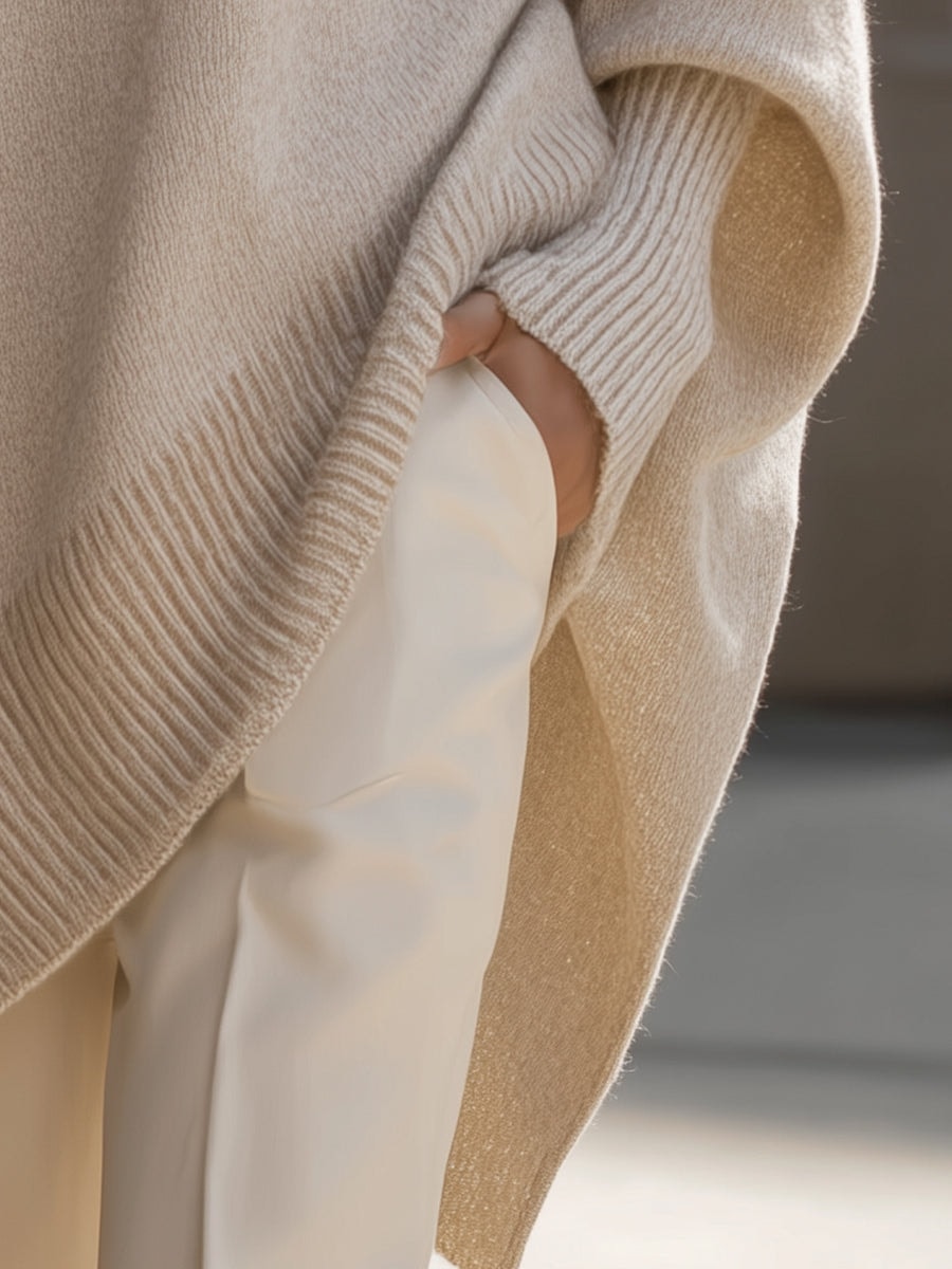 Close side view of the cream poncho sweater showing the extended asymmetric hem over light trousers.