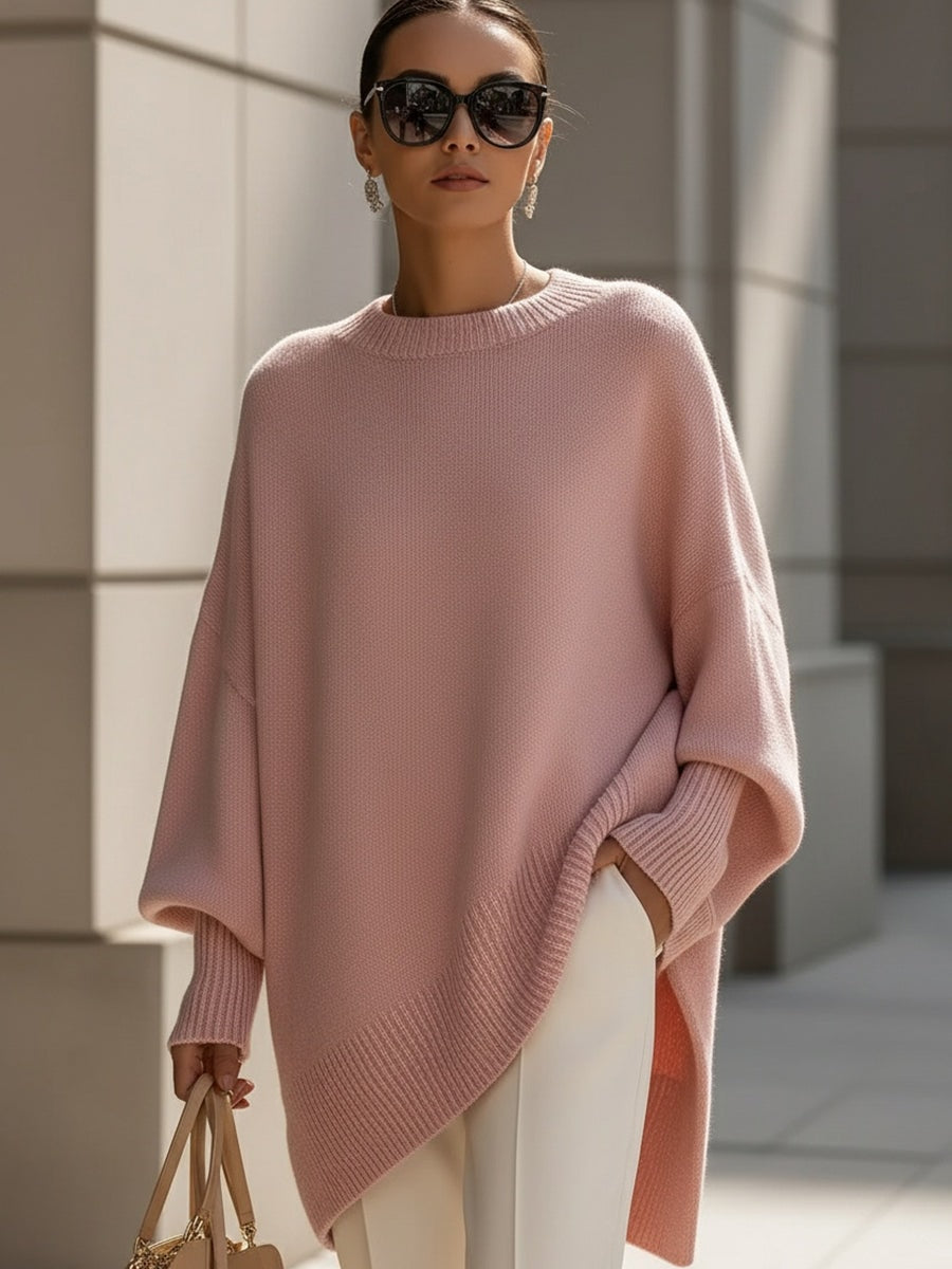 Blush pink asymmetric poncho sweater with ribbed trim, wide sleeves, and a pointed front hem styled with light trousers.