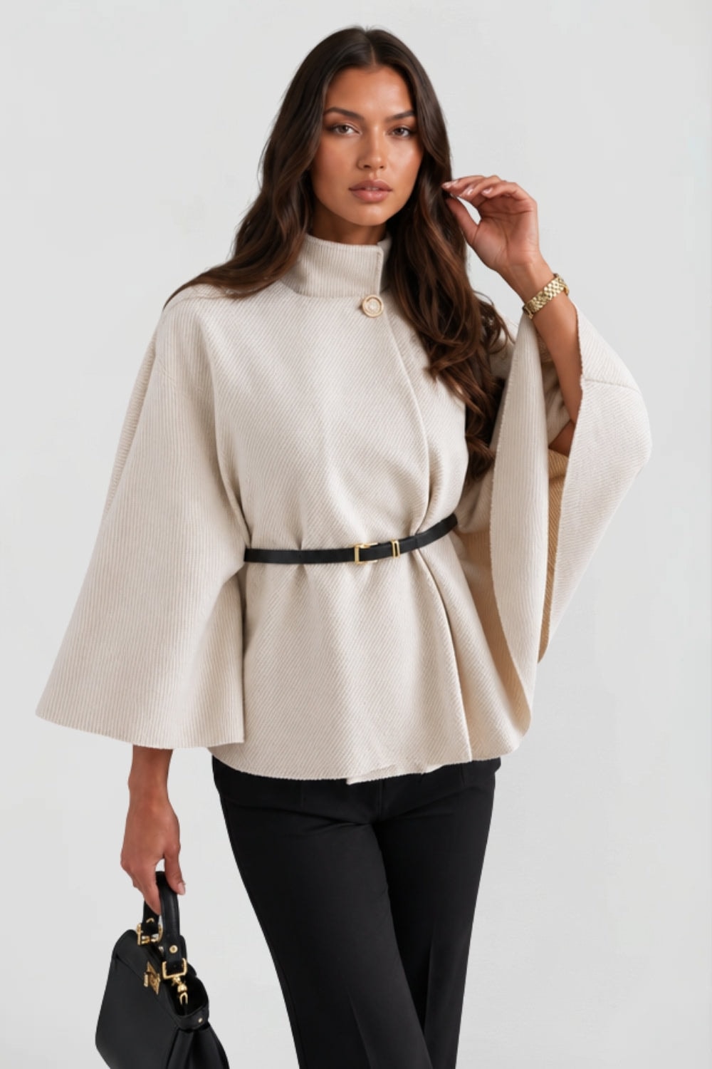 Angled front view of the cream cape jacket with one sleeve raised, revealing the open drape and belted waist.