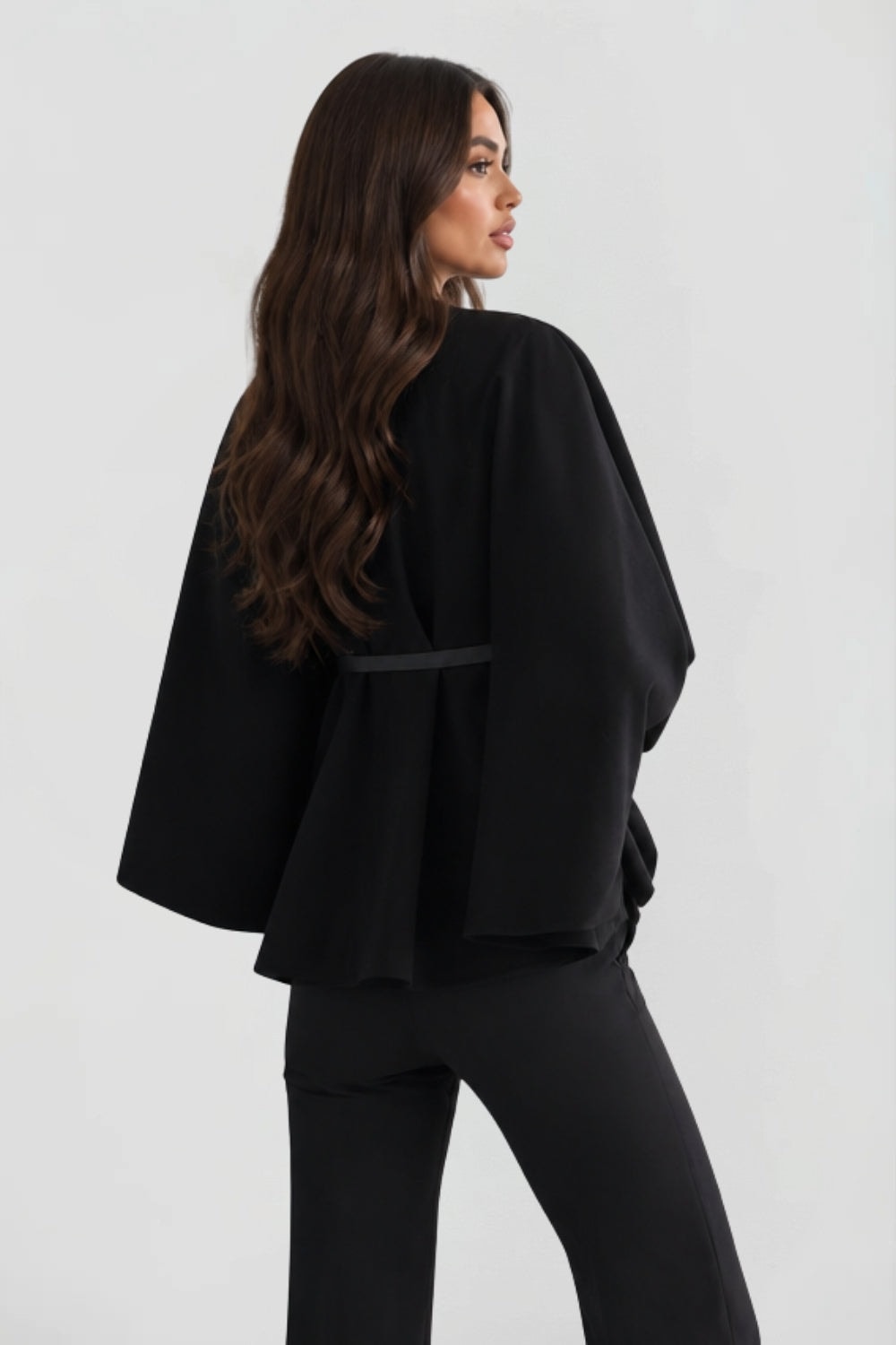 Back three-quarter view of the black cape jacket, emphasizing the flowing back panels and belt.