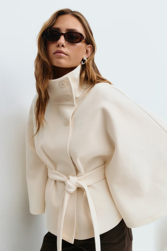 Cream belted cape jacket worn with a high stand collar, front buttons, and wide cape sleeves.
