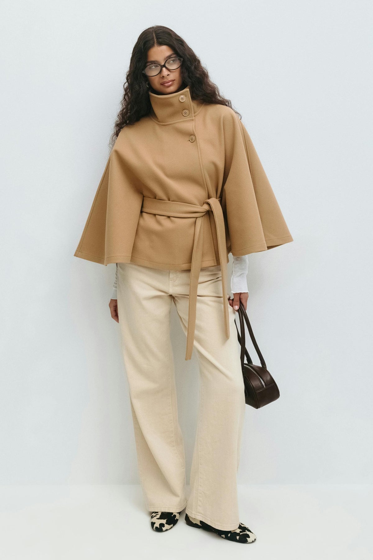 Full-length camel cape jacket outfit with tied waist belt, cream trousers, and a handbag.