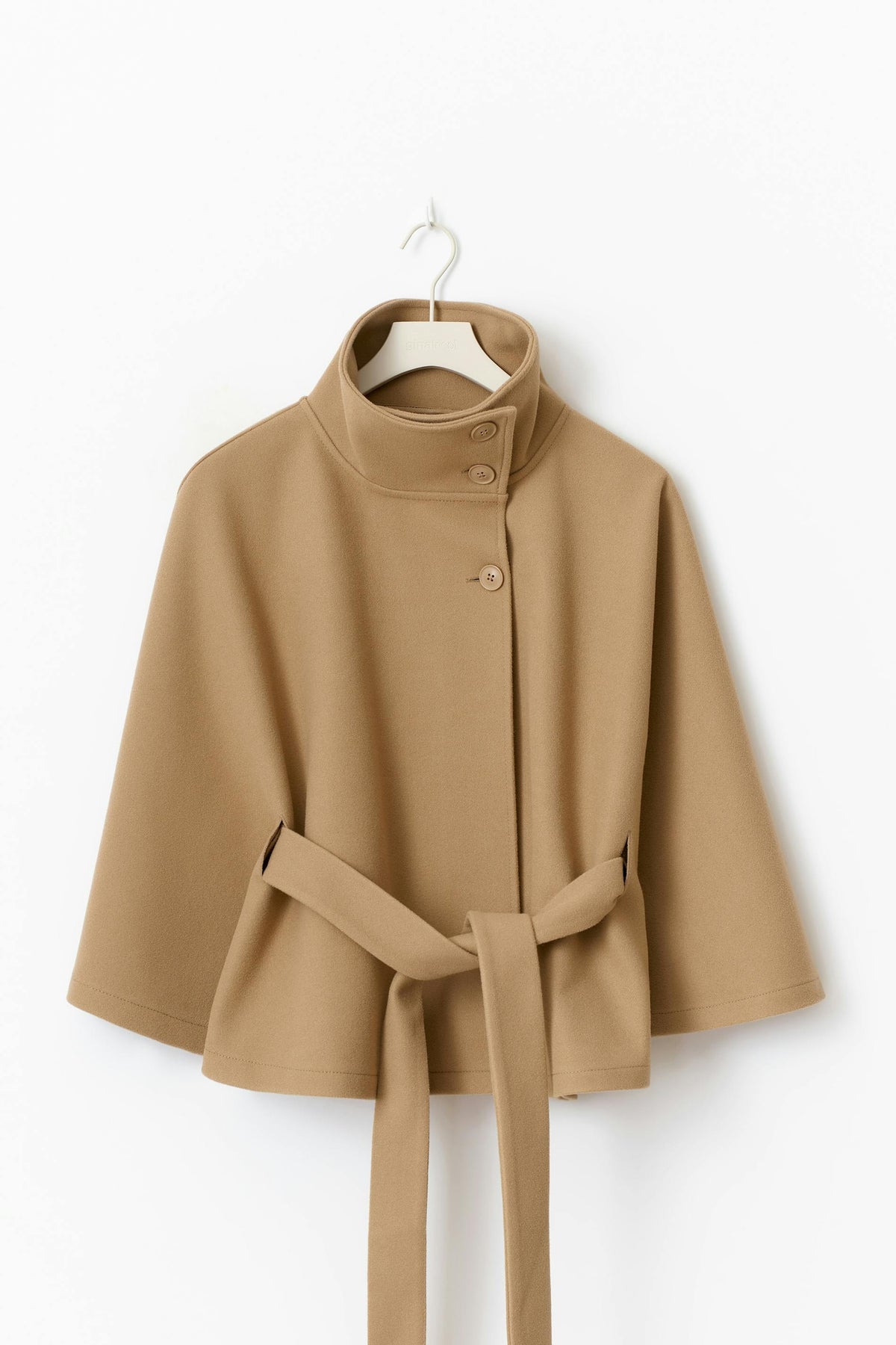 Camel cape jacket displayed on a hanger, showing the raised collar, front button closure, wide sleeves, and belt.