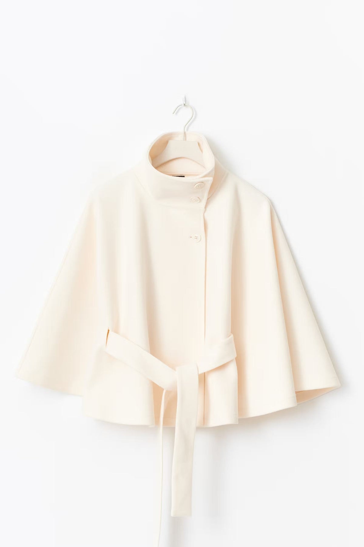 Cream cape jacket displayed on a hanger, showing its short length, stand collar, button front, and tied belt.