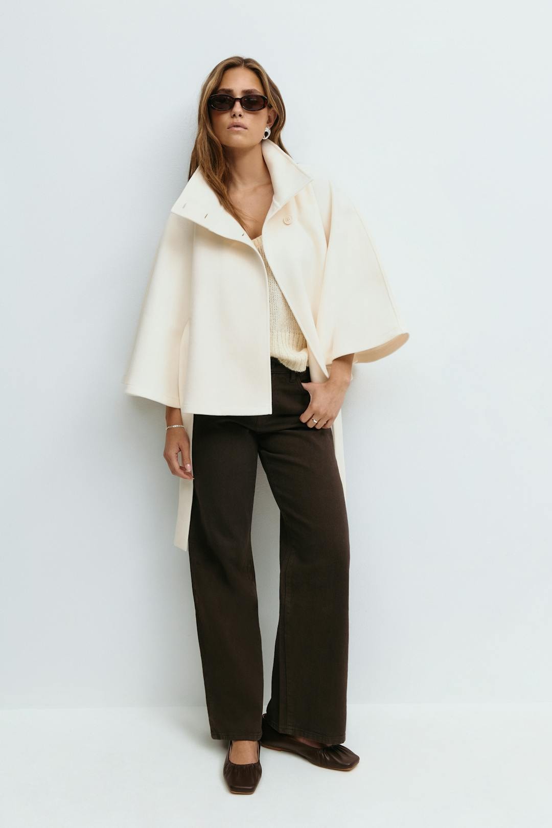 Full-length styling of the cream cape jacket worn open over a light top with trousers.