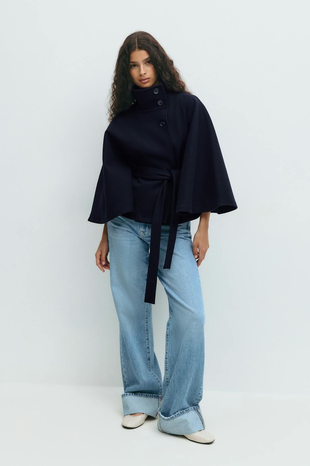 Navy belted cape jacket styled with jeans, showing its short silhouette and dramatic sleeves.
