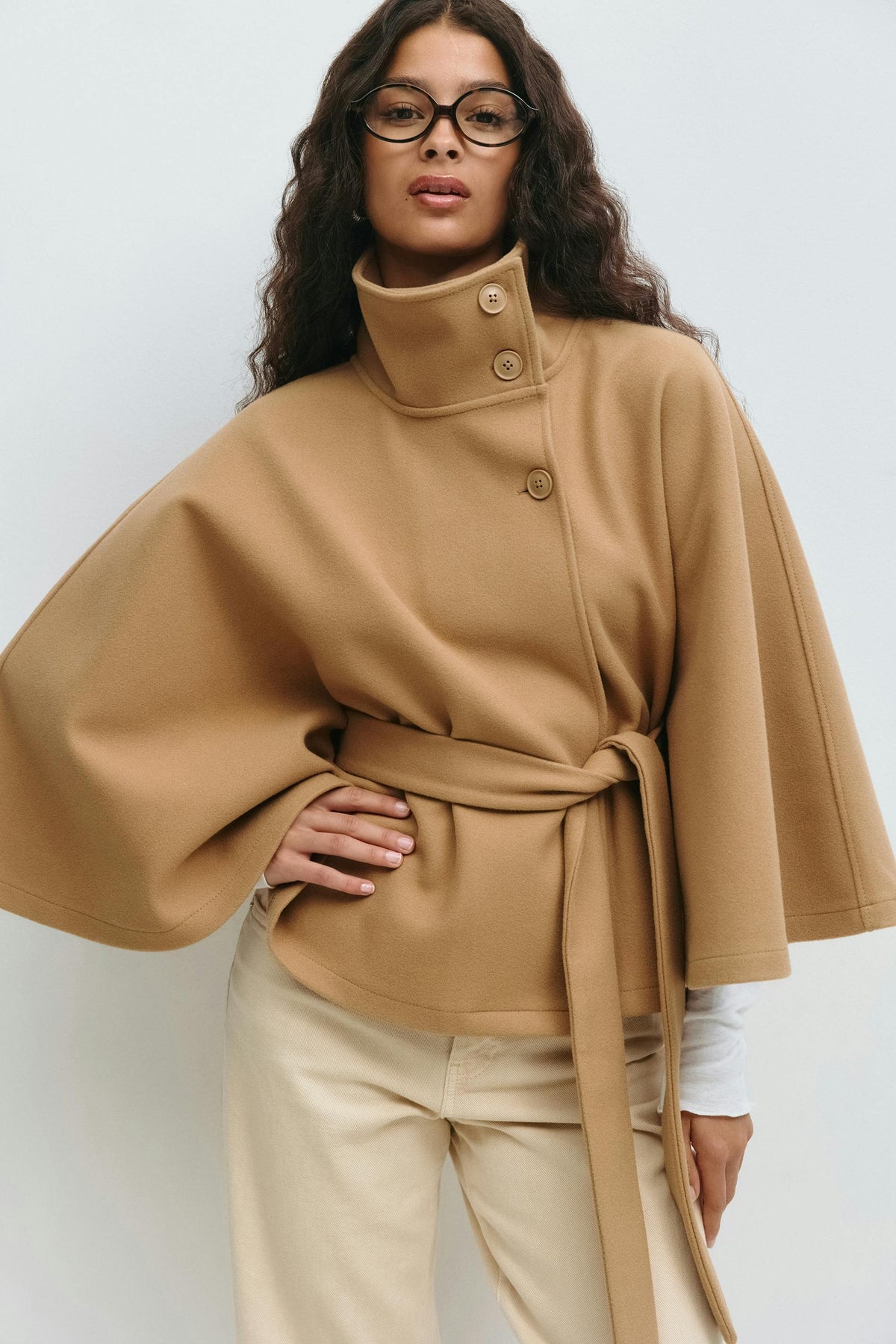 Camel cape jacket worn with a high stand collar, visible front buttons, wide sleeves, and a knotted waist belt.