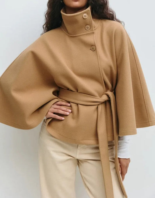 Camel belted high-neck cape coat shown from the front with a buttoned collar and wide sleeves over light trousers.