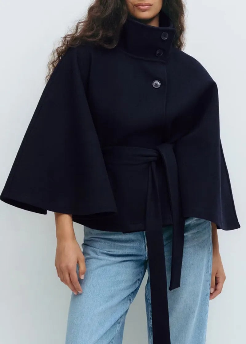 Navy high-neck cape coat styled with jeans, featuring a buttoned collar, flowing cape sleeves, and a long self-tie belt.