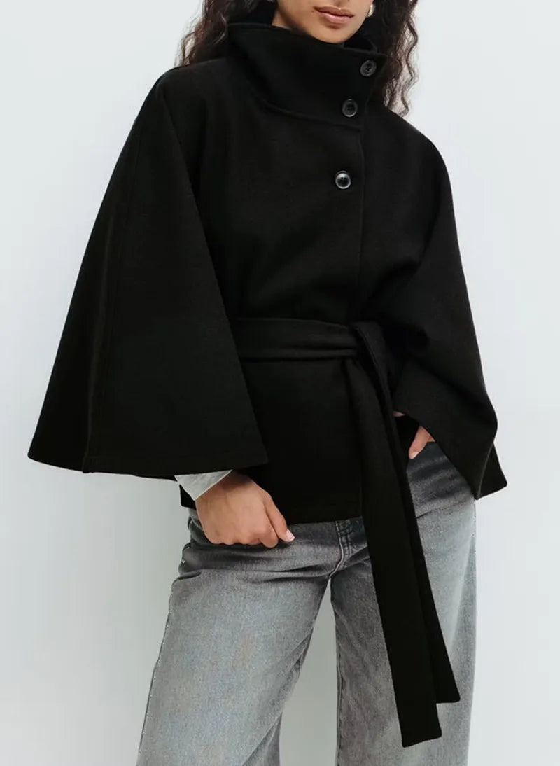 Black high-neck cape coat worn with gray jeans, showing the buttoned collar, relaxed cape sleeves, and hanging waist ties.