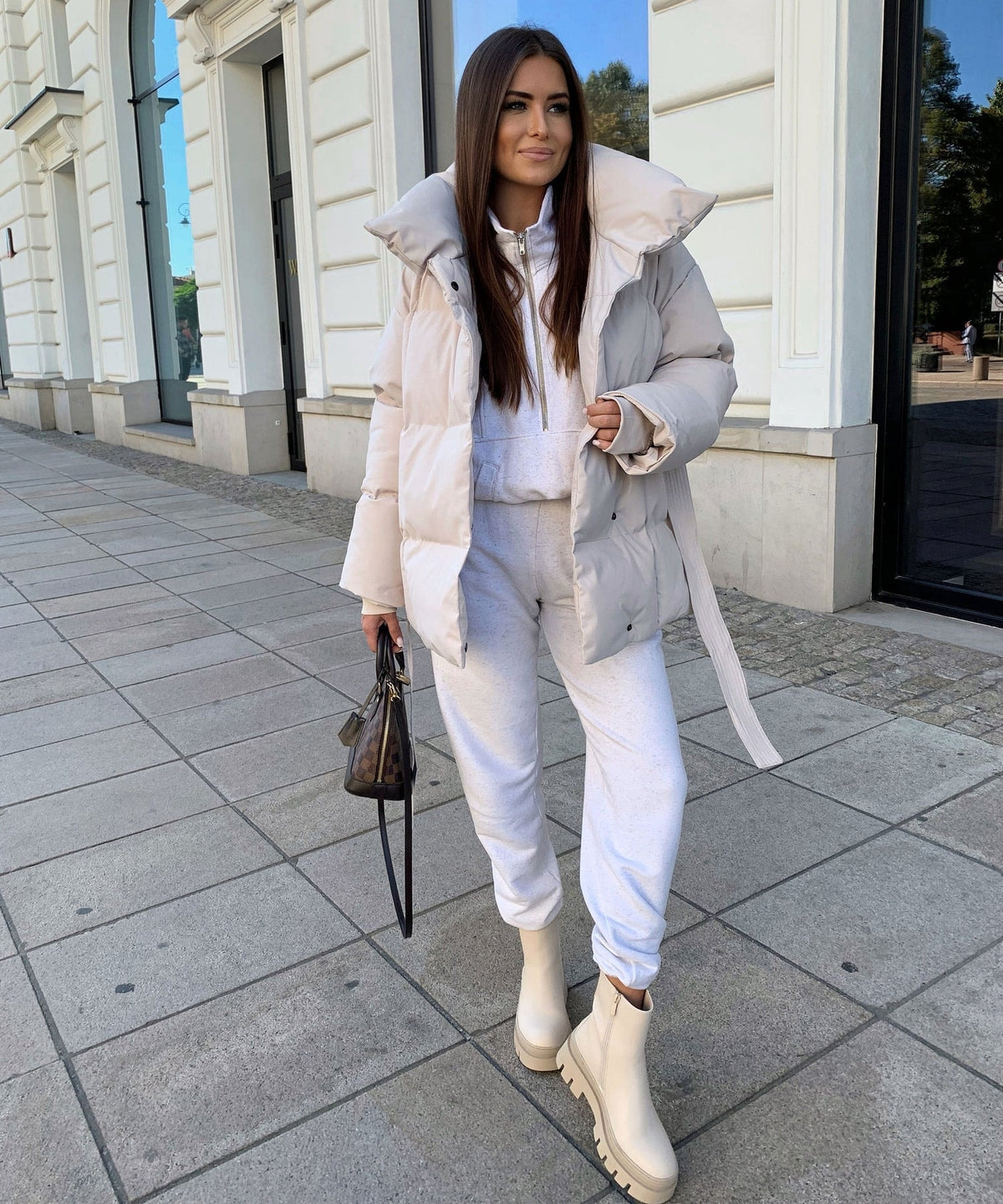 Full-length view of the light beige belted puffer jacket worn open, with the waist tie hanging loosely beside white trousers and ankle boots.