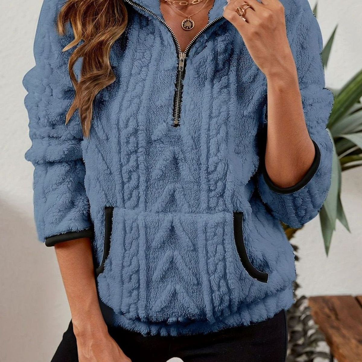 Close front view of the pullover in dusty blue, showing its raised cable texture, stand collar, and contrasting pocket and cuff trim.