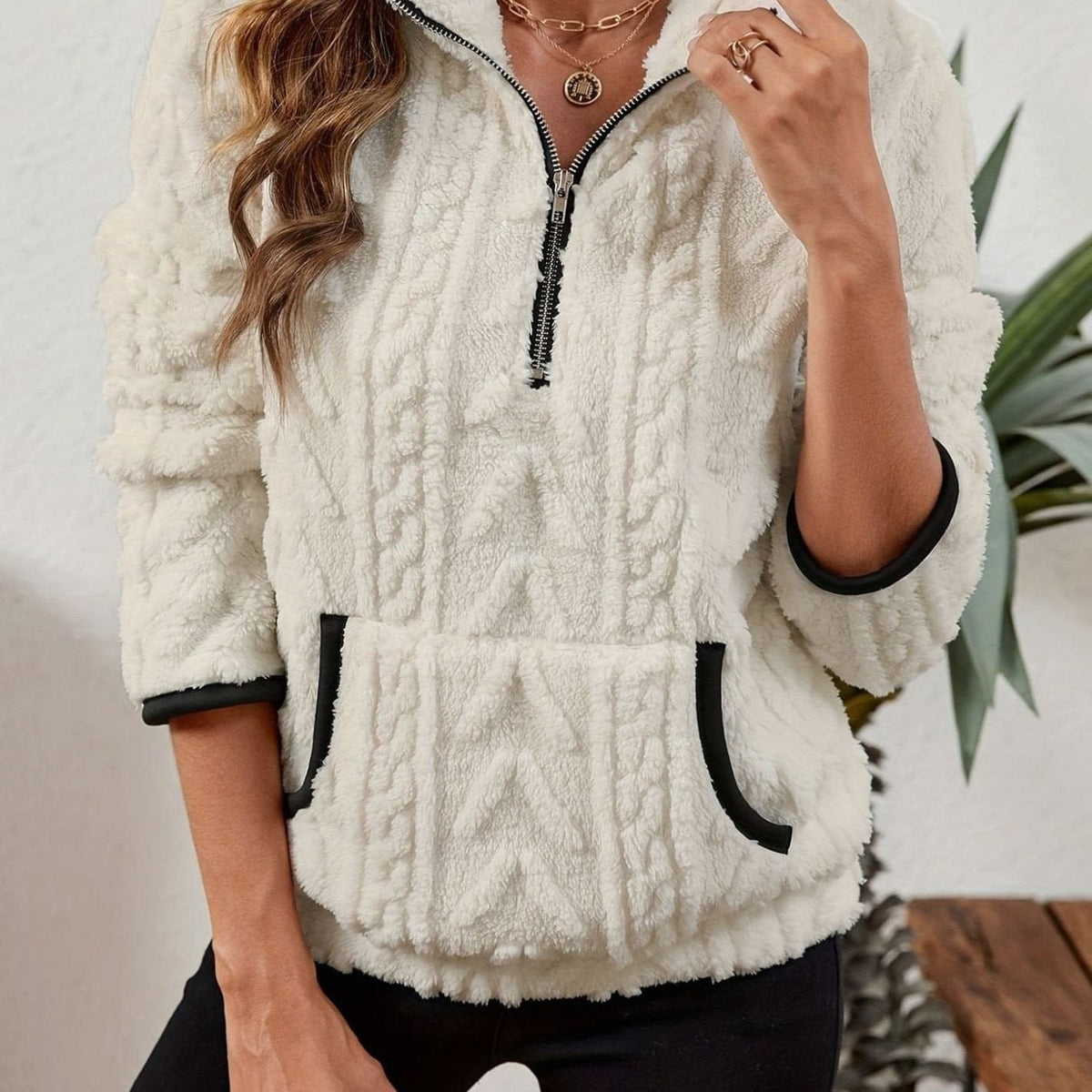 Close front view of the pullover in cream, highlighting the tactile cable texture and dark edging around the pocket and cuffs.