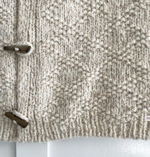Close-up of the vest’s chunky patterned knit and ribbed lower hem, with two wooden toggle closures partly visible.