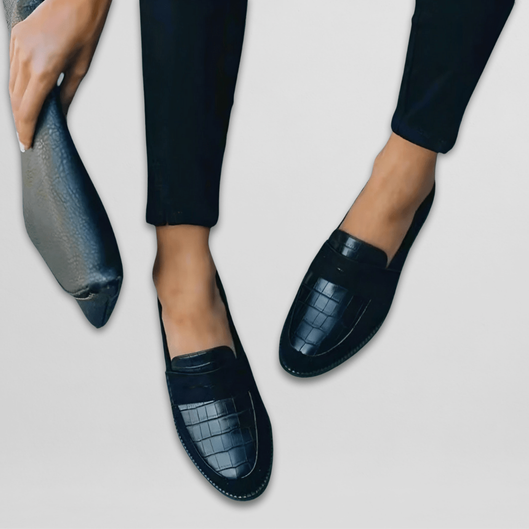 Black croc-embossed loafer pair shown at an offset angle, with one shoe held to reveal its flexible-looking sole and the other worn on foot.