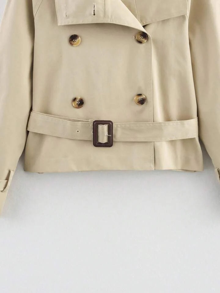 Close-up of the beige cropped trench coat lower front highlighting the double-breasted buttons, belt, buckle, and hem.