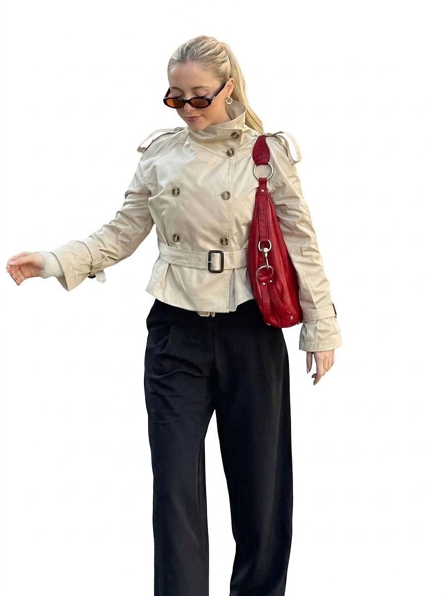 Model wearing the beige cropped trench coat with its belted waist, raised collar, and double-breasted front styled with black trousers.