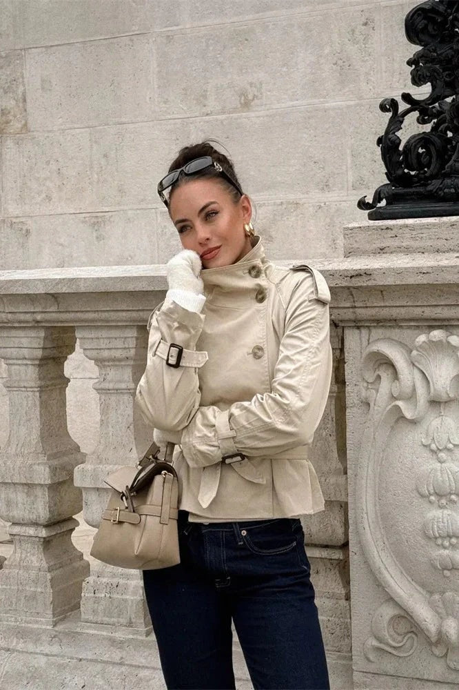 Beige cropped trench coat shown on a model with the waist belt fastened and cuff straps visible while paired with dark jeans.