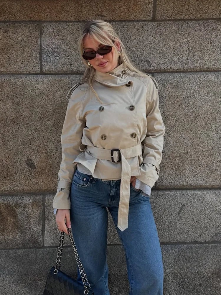 Front view of the belted beige trench coat styled casually with blue jeans, highlighting its cropped hem and structured shoulders.