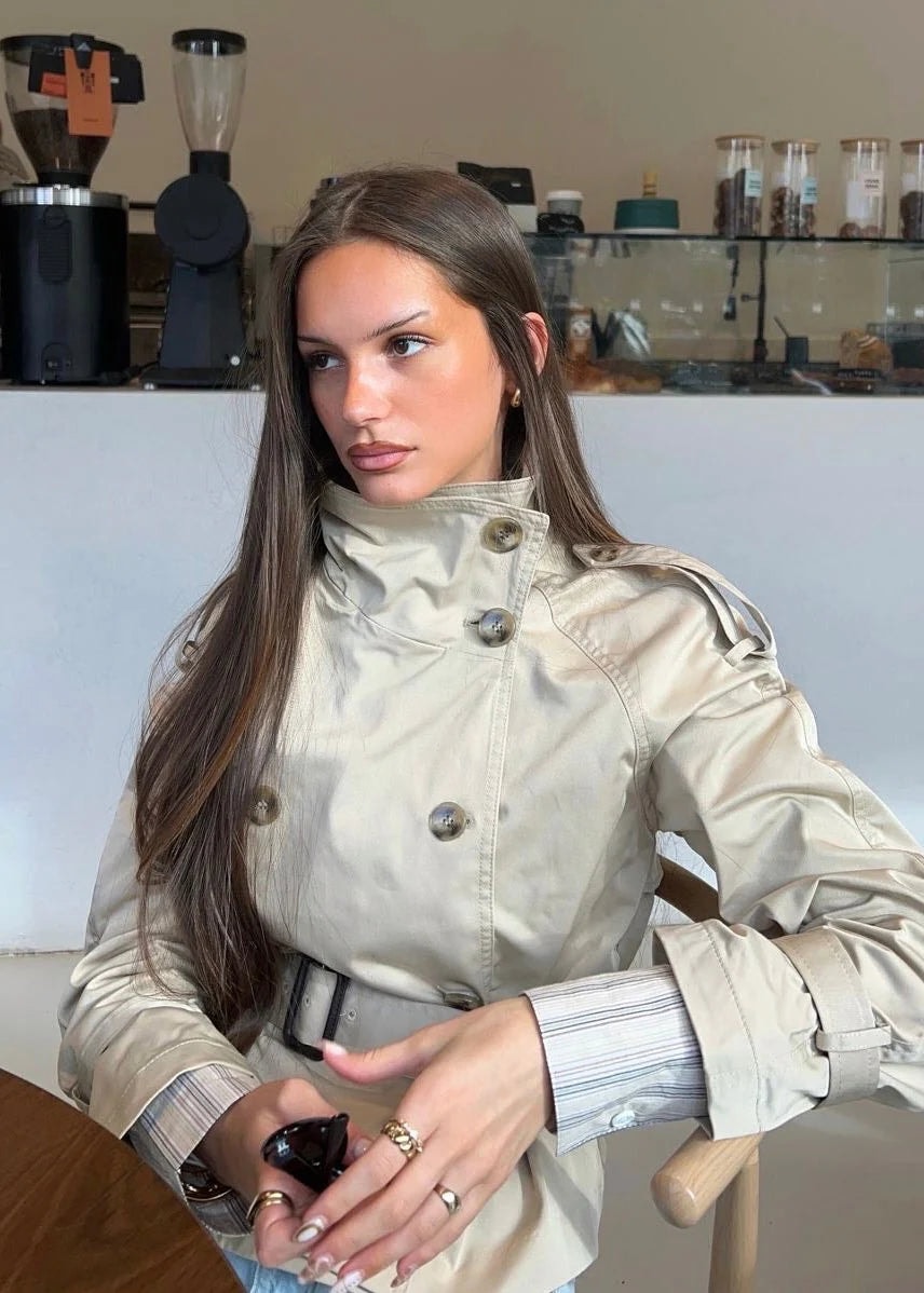 Close view of the beige trench coat showing the stand collar, shoulder epaulettes, double-breasted buttons, and waist belt.