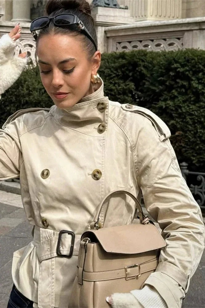 Model wearing the beige double-breasted trench coat with the belt fastened and cuff straps visible at the wrists.