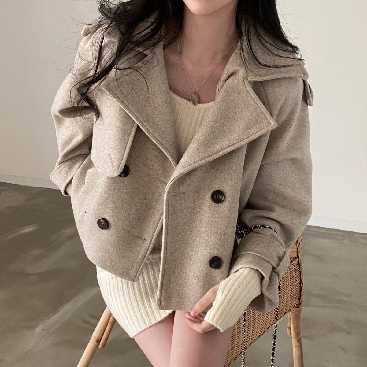 Front view of a taupe cropped double-breasted peacoat with a wide notched collar, dark buttons, long sleeves, side pockets, and tabbed cuffs over a light ribbed sweater.