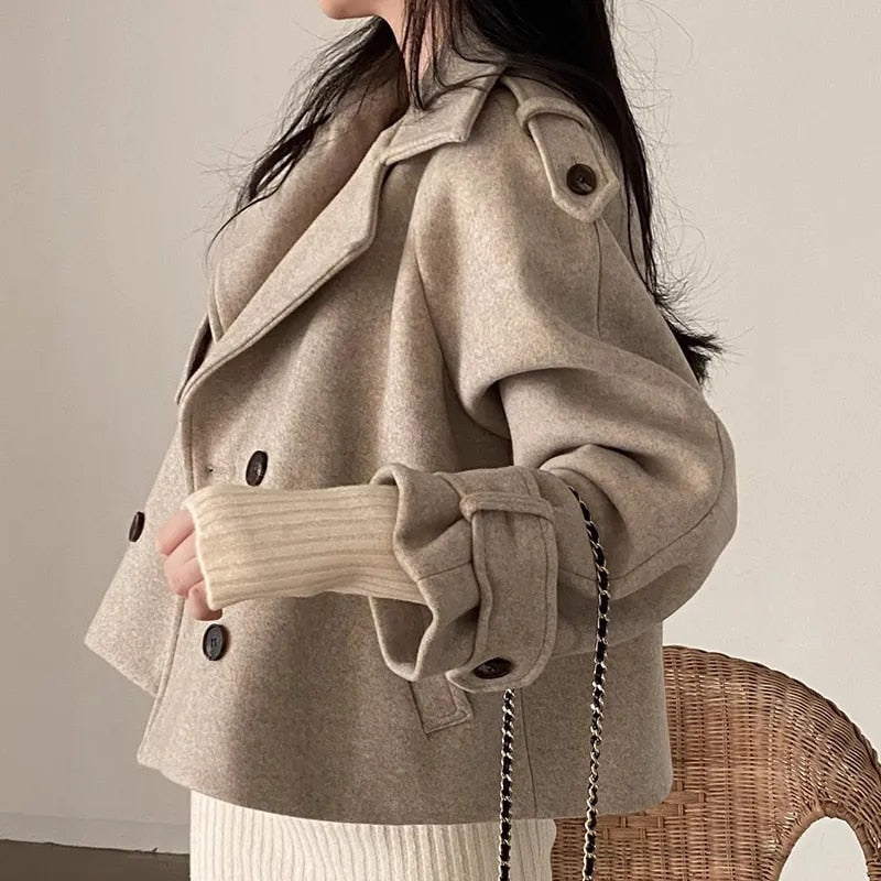 Side view of the taupe cropped peacoat showing its short structured shape, layered notched collar, shoulder tab, long sleeve, and buttoned cuff tab.