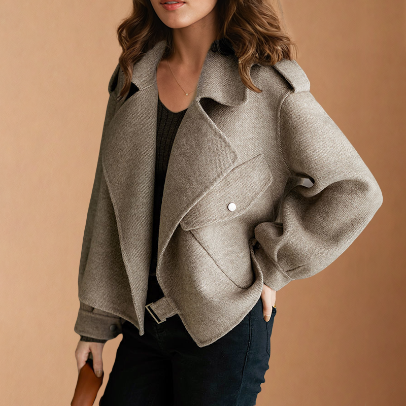 Cropped jacket styled with one hand in a pocket, highlighting the wide lapels, shoulder epaulettes, flap pocket, buttoned cuff, and front hem buckle.