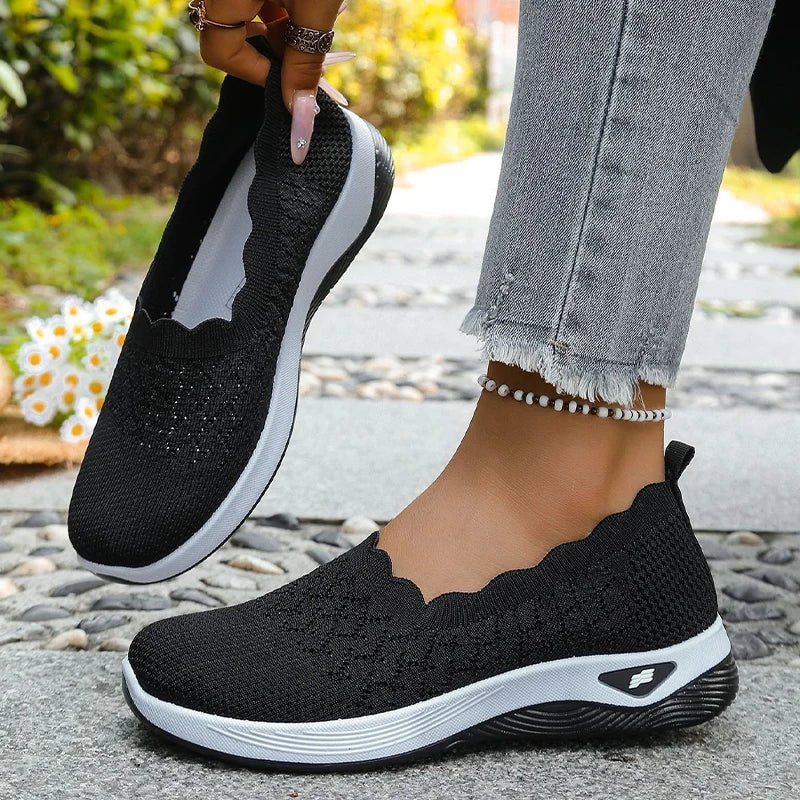 Black knit slip-on walking shoes shown in close-up with white cushioned soles and scalloped openings.