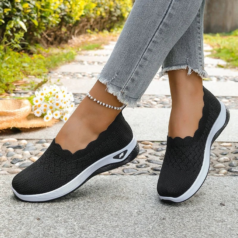 Black knit slip-on shoes worn on both feet, viewed at an angled walking stance outdoors.