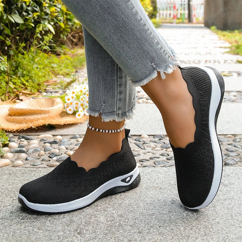 Black knit slip-on shoes shown with one foot grounded and the other lifted to reveal the side and sole.