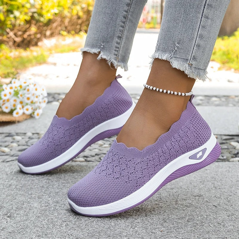 Purple knit slip-on walking shoes worn on both feet, showing scalloped openings and matching cushioned soles.