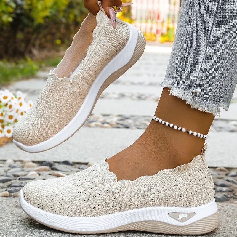 Beige knit slip-on shoe lifted by the heel beside another shoe, showing the side profile and flexible upper.