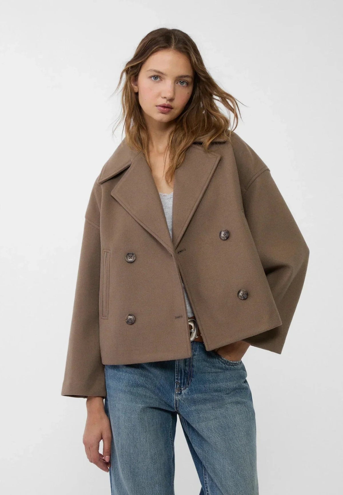 Model in the brown cropped jacket styled with blue jeans, highlighting its angled front closure and slanted pockets.