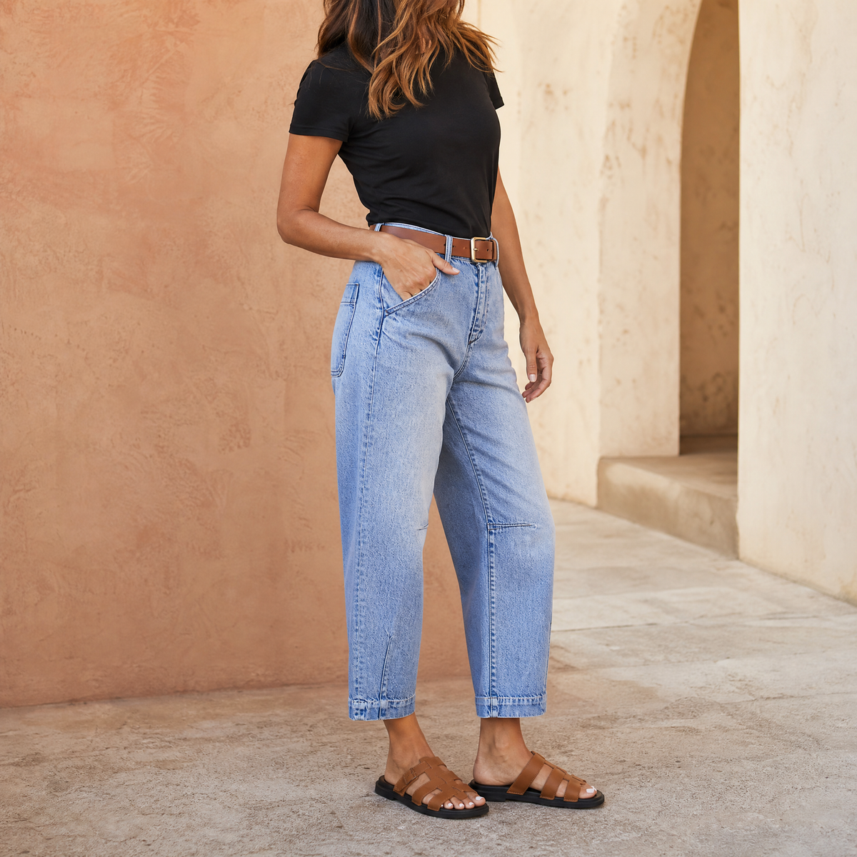 Model wearing light-blue cropped barrel-leg jeans with a high waist, front pockets, and relaxed curved legs styled with a black top and flat sandals.