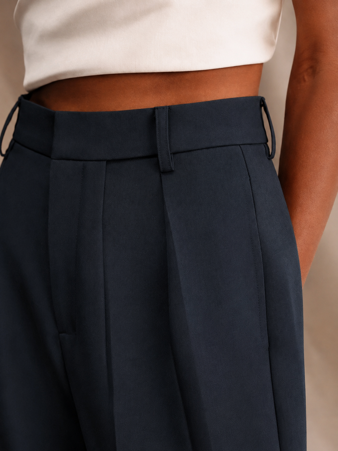 Close-up of navy trousers showing the high waistband, belt loops, front pleat, and side pocket.