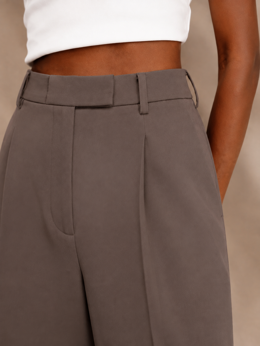Close-up of taupe trousers showing the high waistband, front pleat, belt loops, and pocket detail.