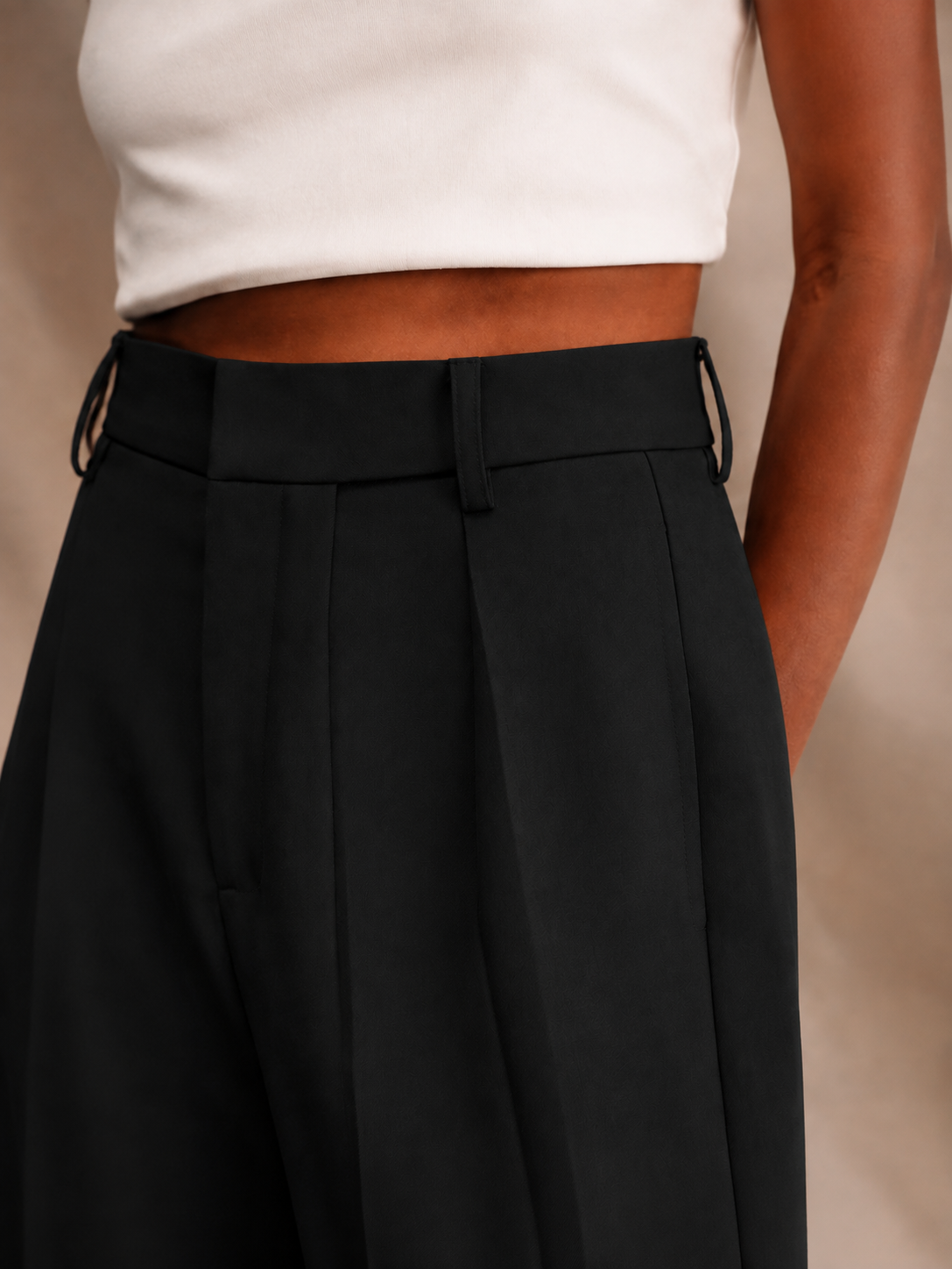 Close-up of black trousers with a high waistband, belt loops, front pleats, and a visible side pocket.
