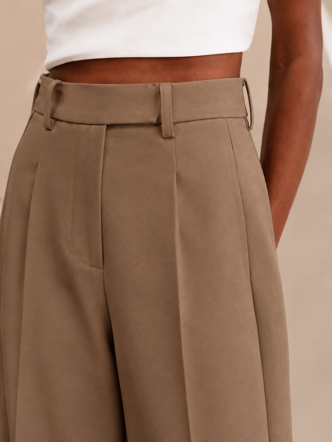Close-up of brown trousers showing the waistband, center belt loop, belt loops at the sides, and tailored pleats.