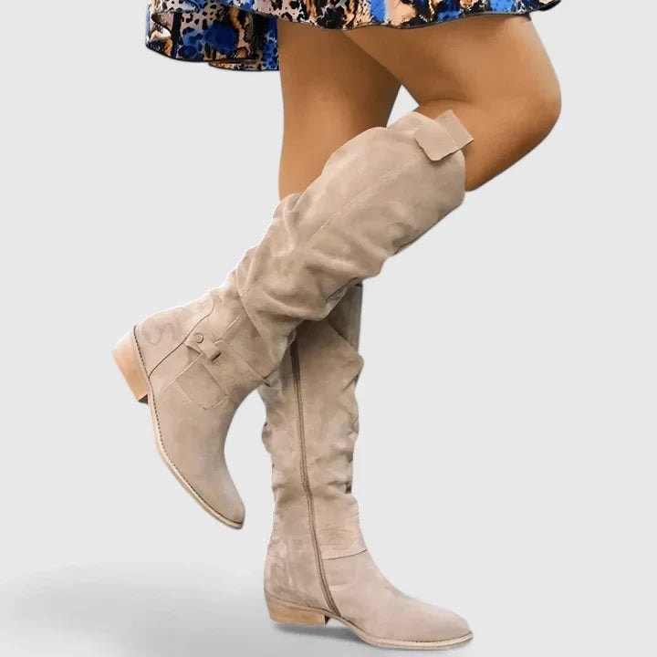 Light beige knee-high boots with ruched shafts, rounded toes, ankle buckle details, side zips, and low heels shown in motion beneath a patterned skirt.