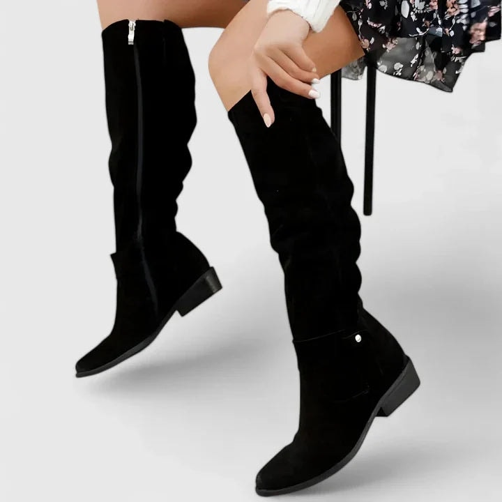 Black knee-high boots with smooth shafts, rounded toes, visible side zips, ankle buckle details, and low block heels worn with a floral skirt.