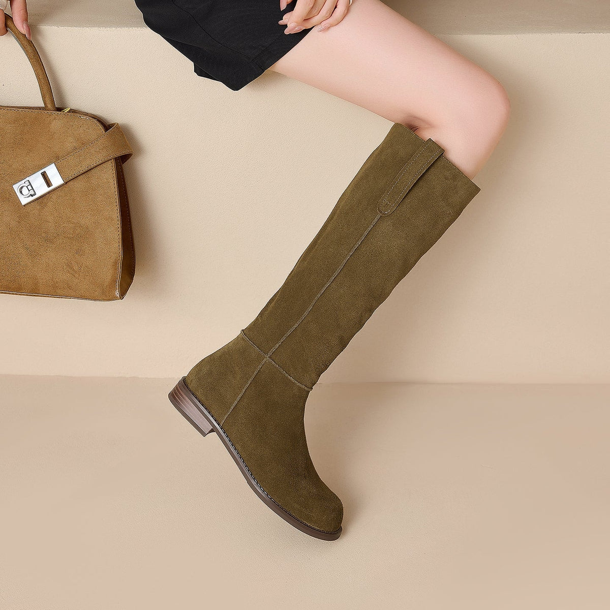 Top-down view of an olive-tan knee-high boot beside a handbag, showing the shaft seam, pull tab, rounded toe, and stacked heel.