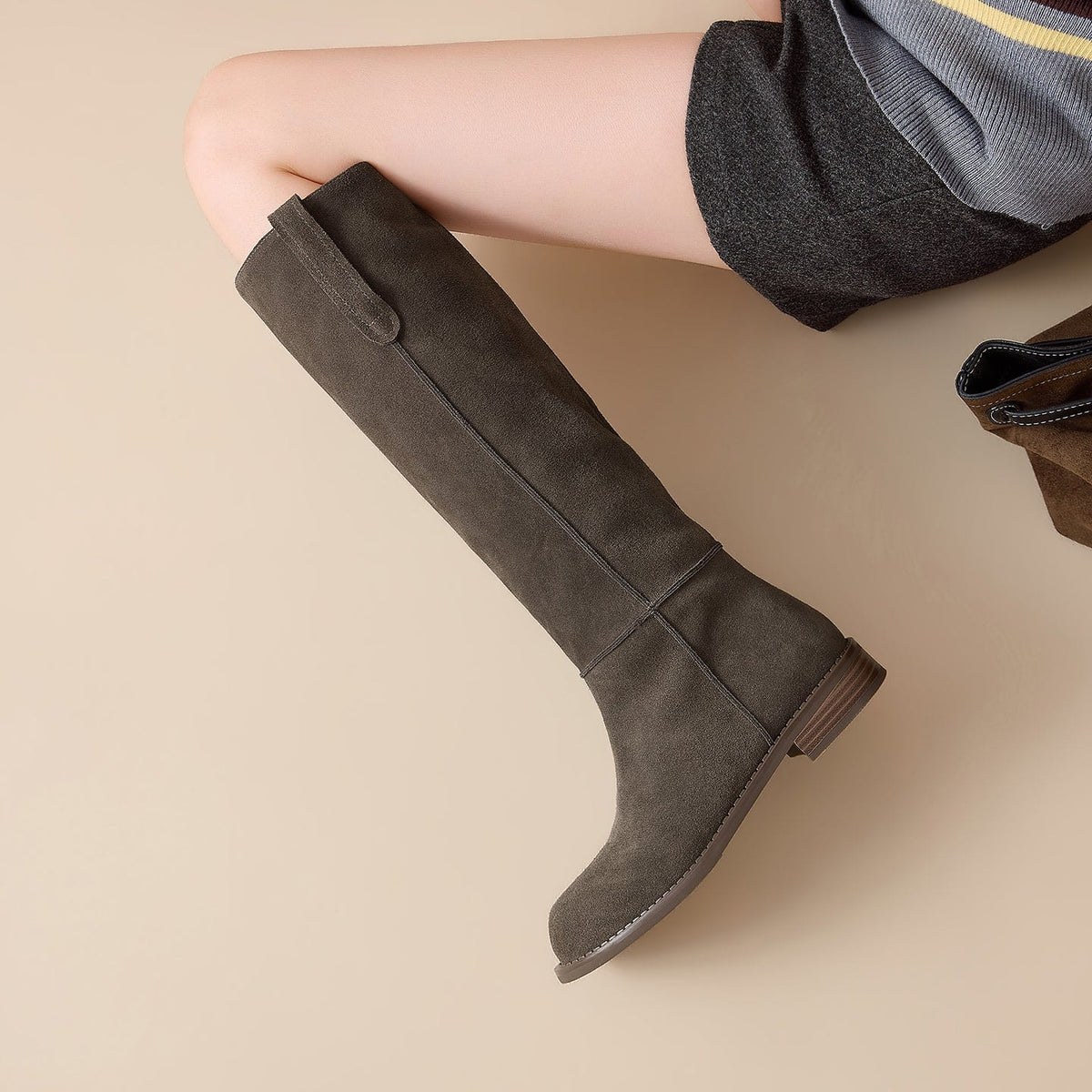 Overhead side view of a charcoal gray knee-high boot worn with a skirt, highlighting its rounded toe, long seam, pull tab, and stacked heel.