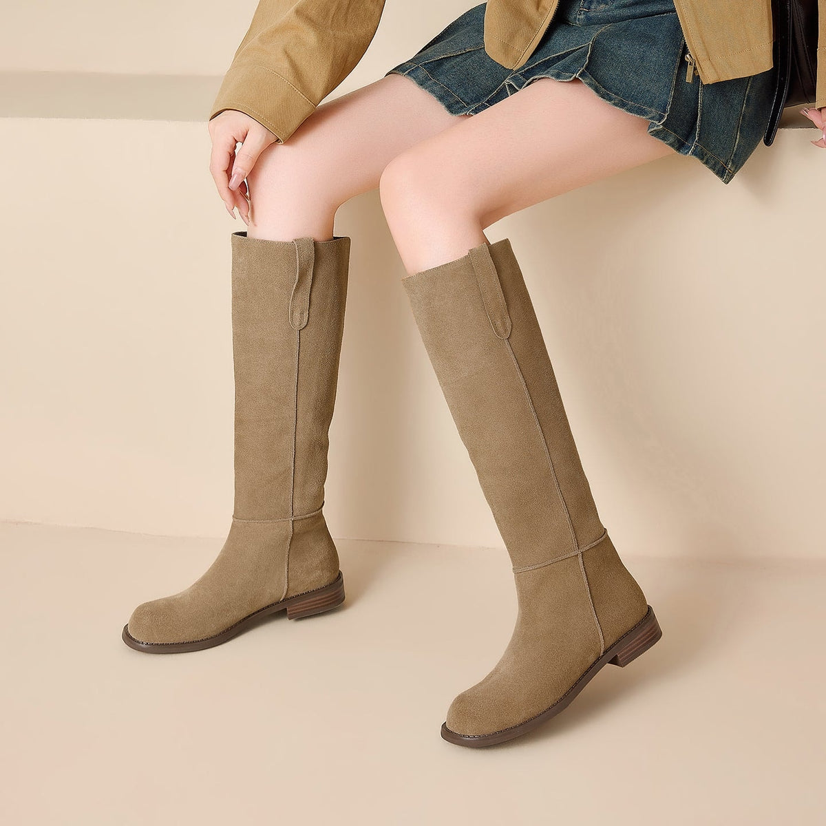 Tan knee-high boots styled with a denim skirt, showing their streamlined shafts and low heels from a seated angle.