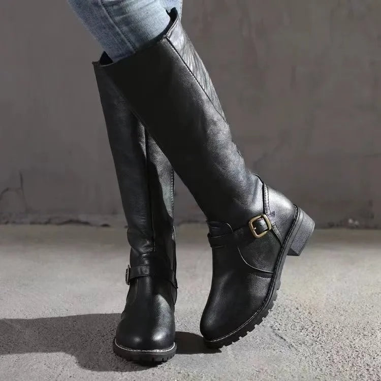 Black knee-high riding boots shown in a crossed-leg pose, with ankle buckle straps, rounded toes, and low block heels.