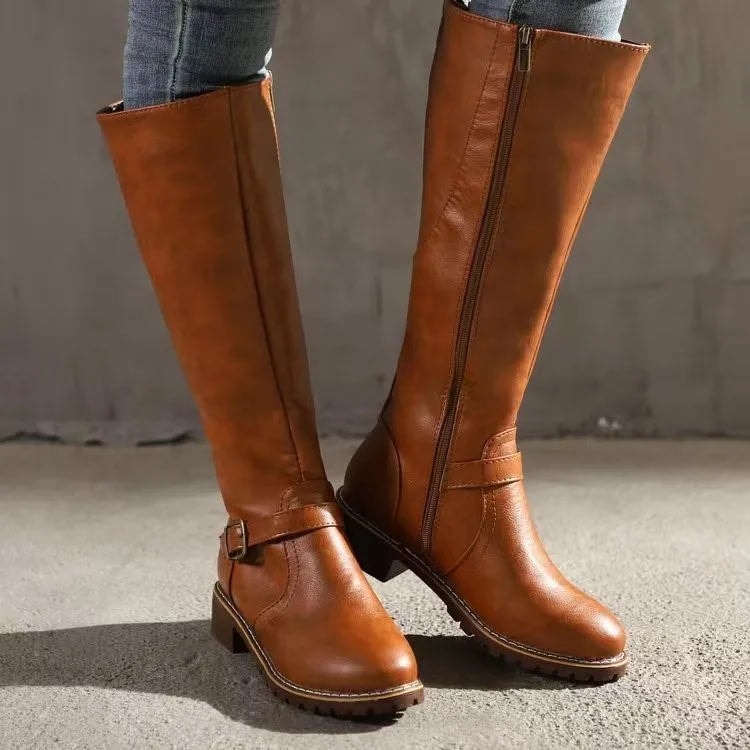 Warm tan knee-high riding boots paired with jeans, featuring rounded toes, ankle buckles, side zippers, and low block heels.