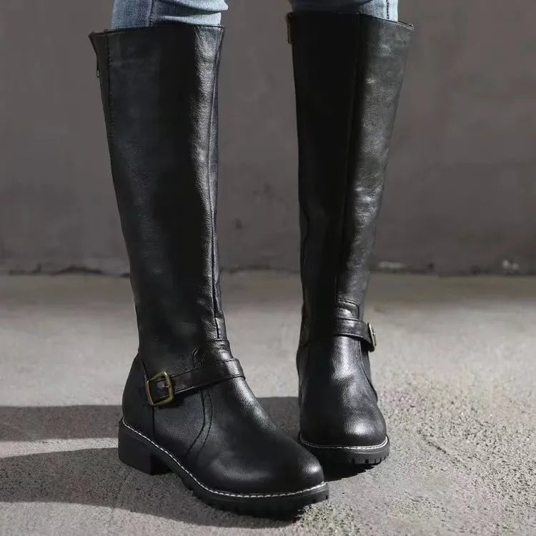 Black knee-high riding boots standing side by side on the ground, clearly showing the front buckle detail, rounded toes, and textured outsoles.